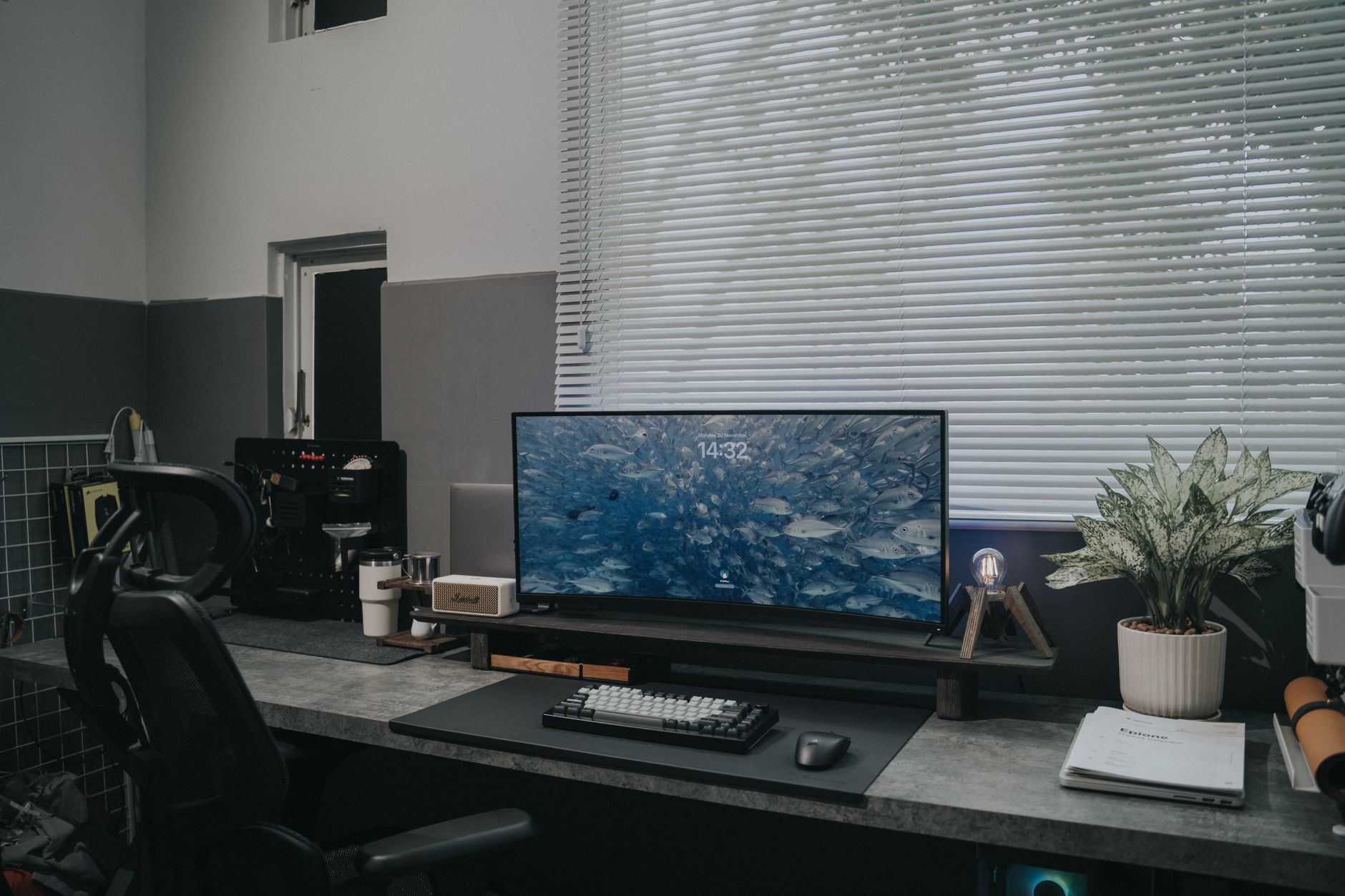 Bureau moderne avec grand écran, clavier et plante verte devant une fenêtre à lattes.
